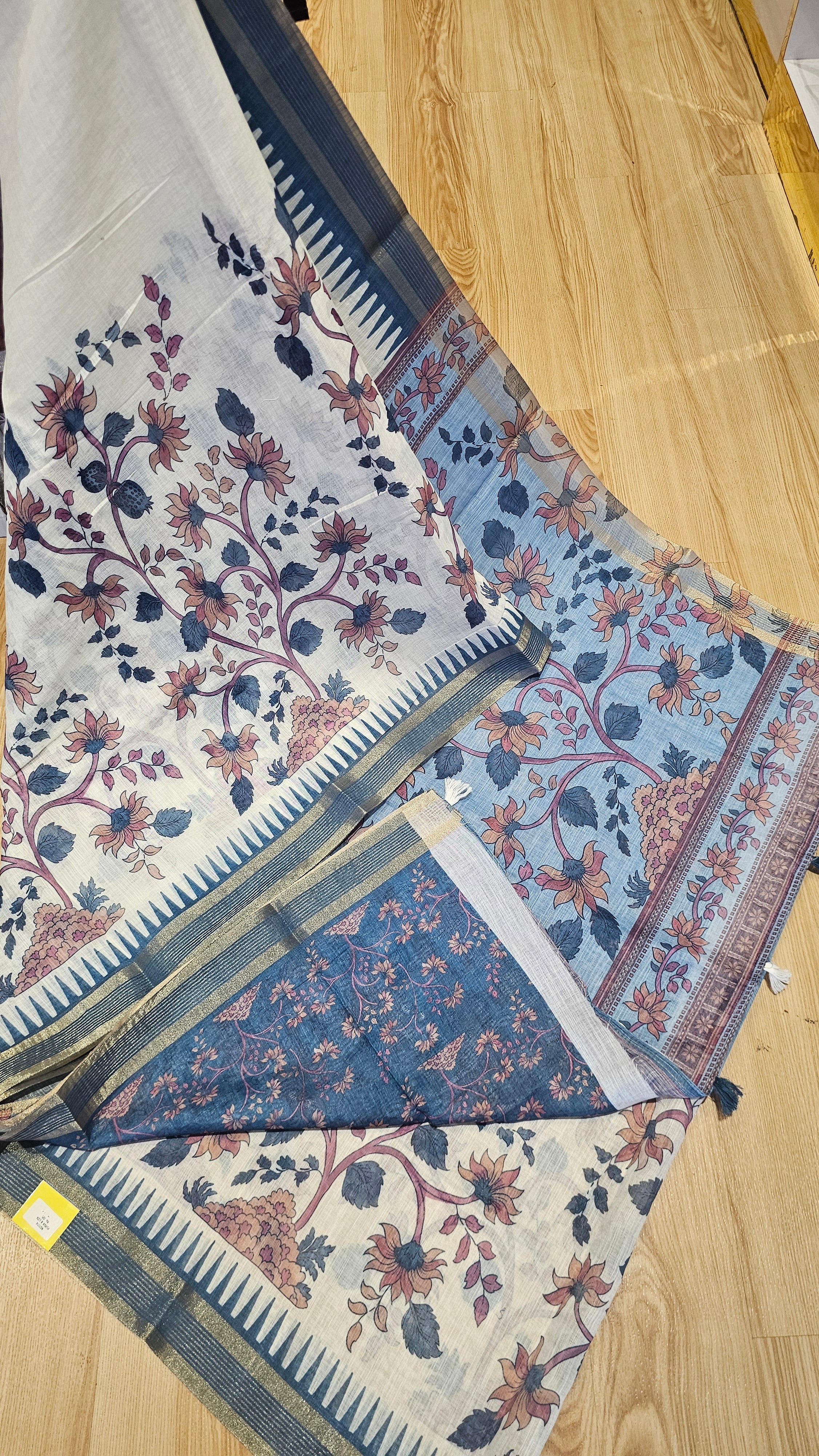 A folded and draped saree with a cream base, featuring a blue and dusty rose floral block print and a blue border, laid out on a light wood floor.