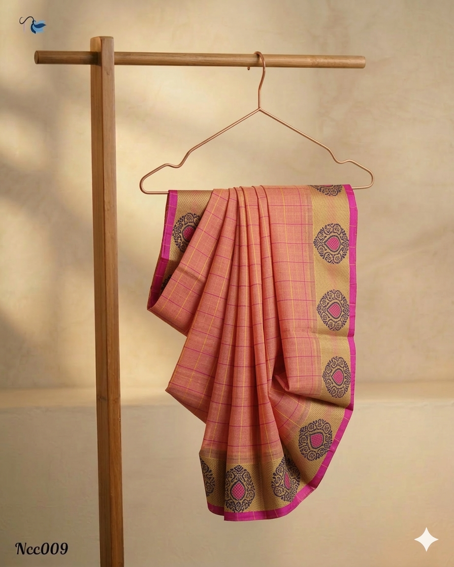 A peach-colored Chettinad cotton saree with small yellow checks hanging on a rose gold hanger. The saree features a wide beige border with purple circular traditional motifs and a thin, bright magenta pink edge.