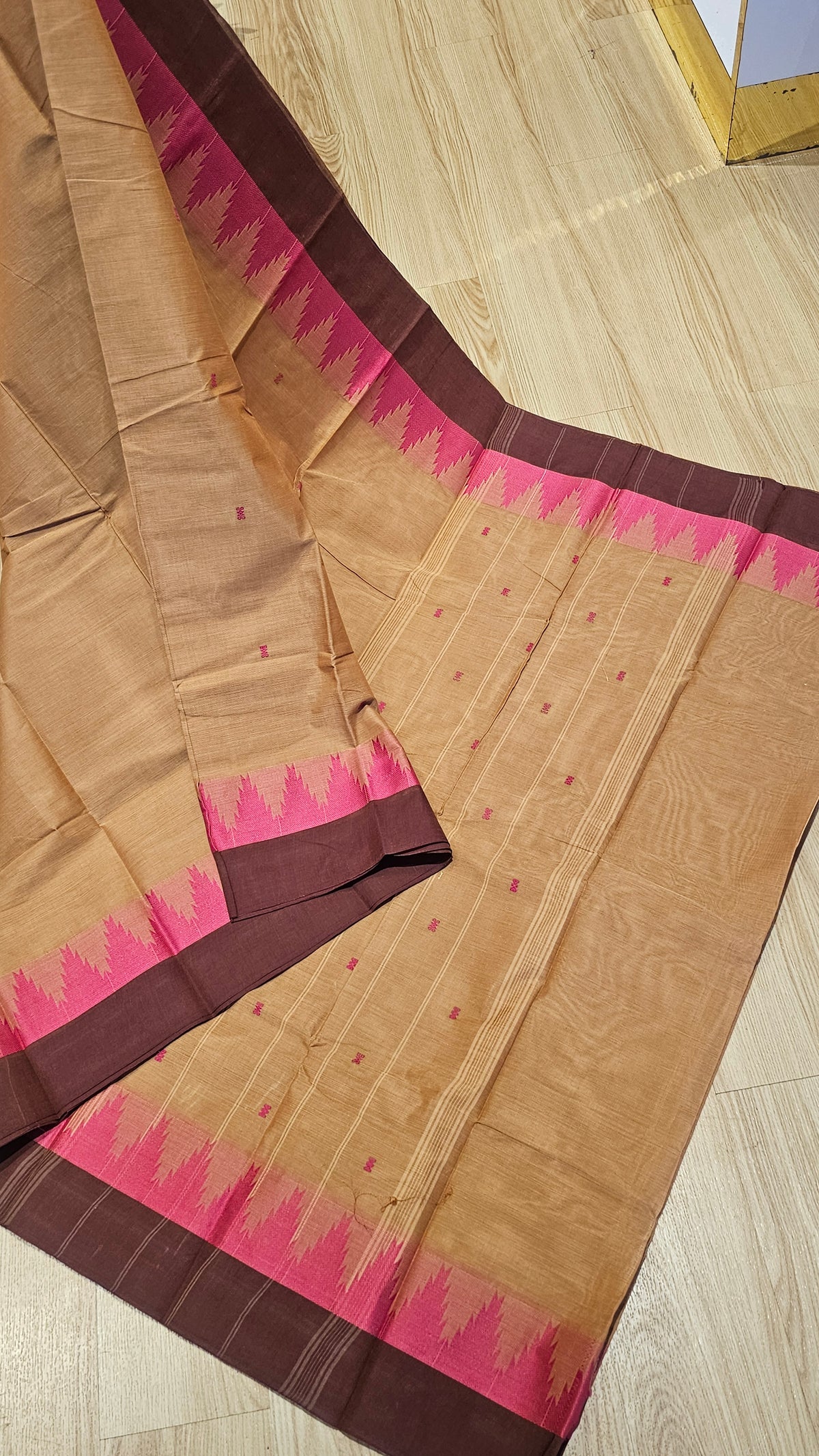 A beige cotton saree with a dark brown border and a vibrant pink and brown zigzag patterned pallu, laid out on a light wood floor.