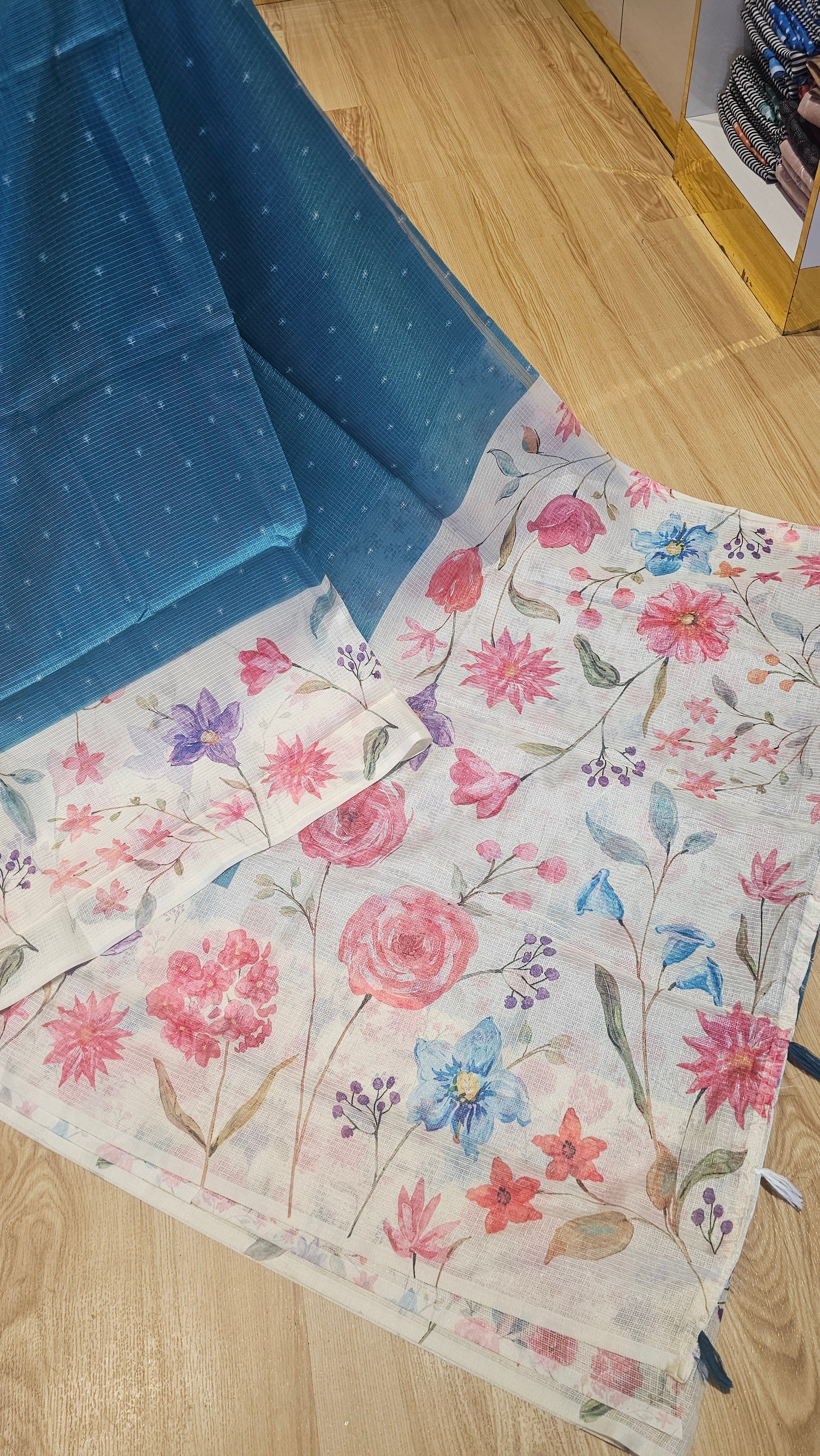 A folded blue dotted fabric garment, likely a sari or scarf, laid next to a white dupatta featuring a pink, blue, and purple floral print, displayed on a light wood floor.