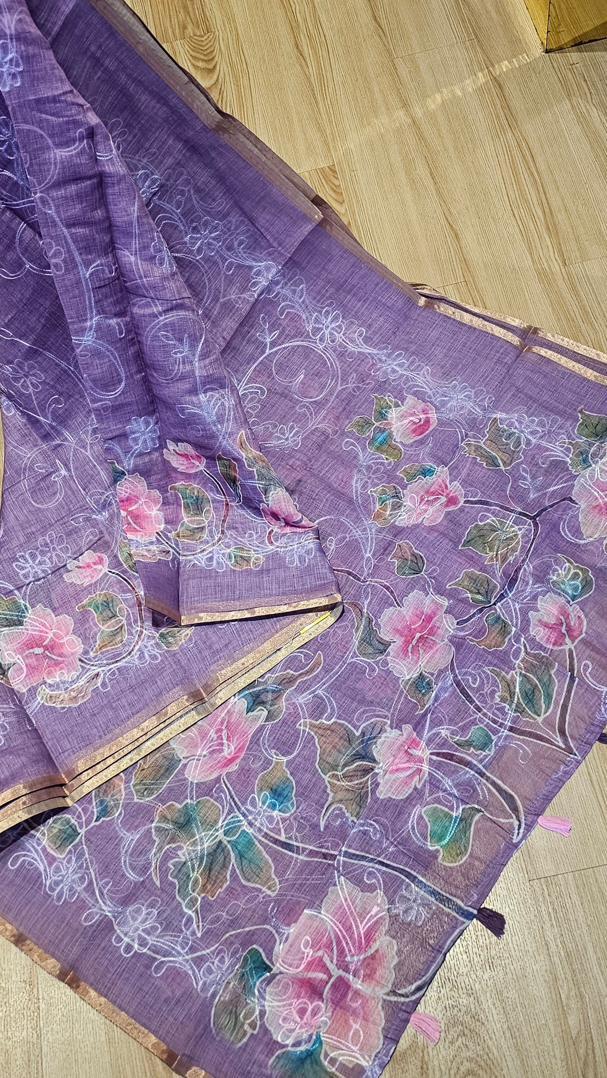 A lavender purple saree with pink floral prints, green leaves, and white vine patterns, laid out on a light wood floor.