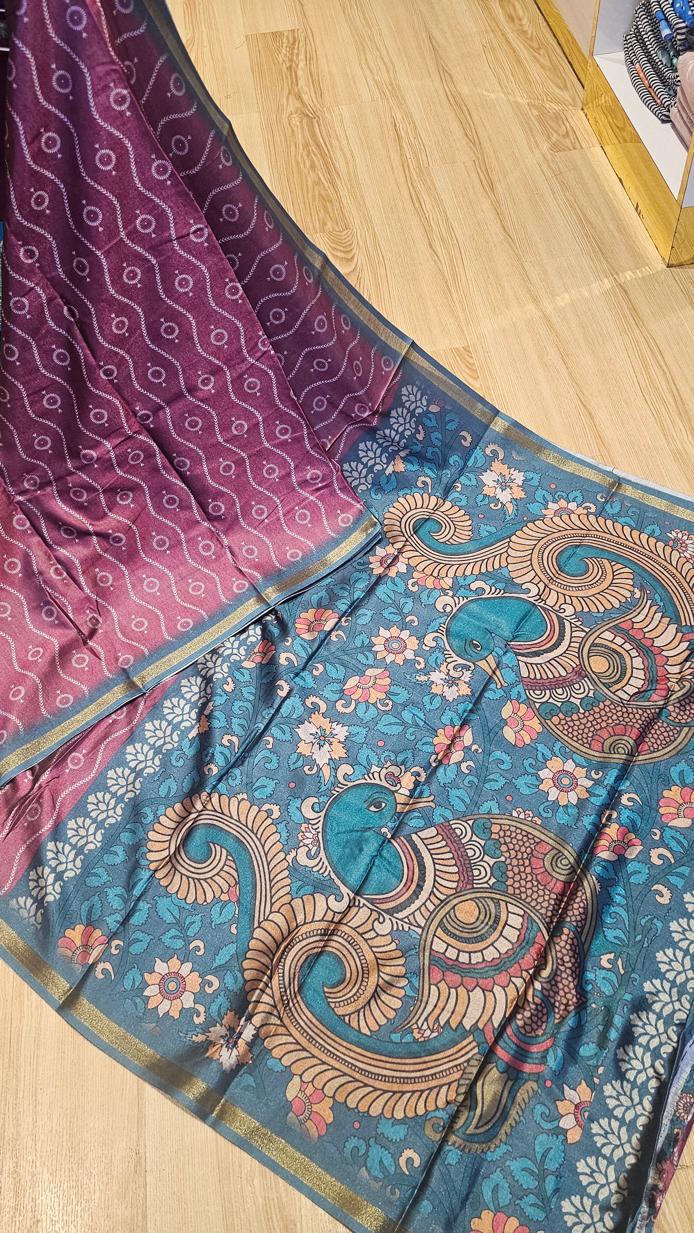 A traditional Indian saree with a teal blue body featuring a large peacock motif and floral patterns, transitioning to a magenta-purple border with a white geometric pattern, laid out on a light wood floor.