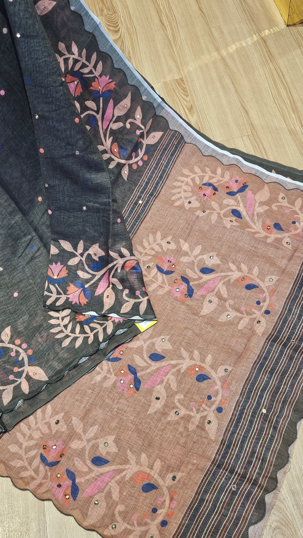 A close-up of a traditional Indian saree or fabric laid flat on a light wood floor. The fabric has a dark charcoal gray border with colorful floral embroidery in pink, blue, and green, and a large dusty pink panel with matching floral motifs and striped details.
