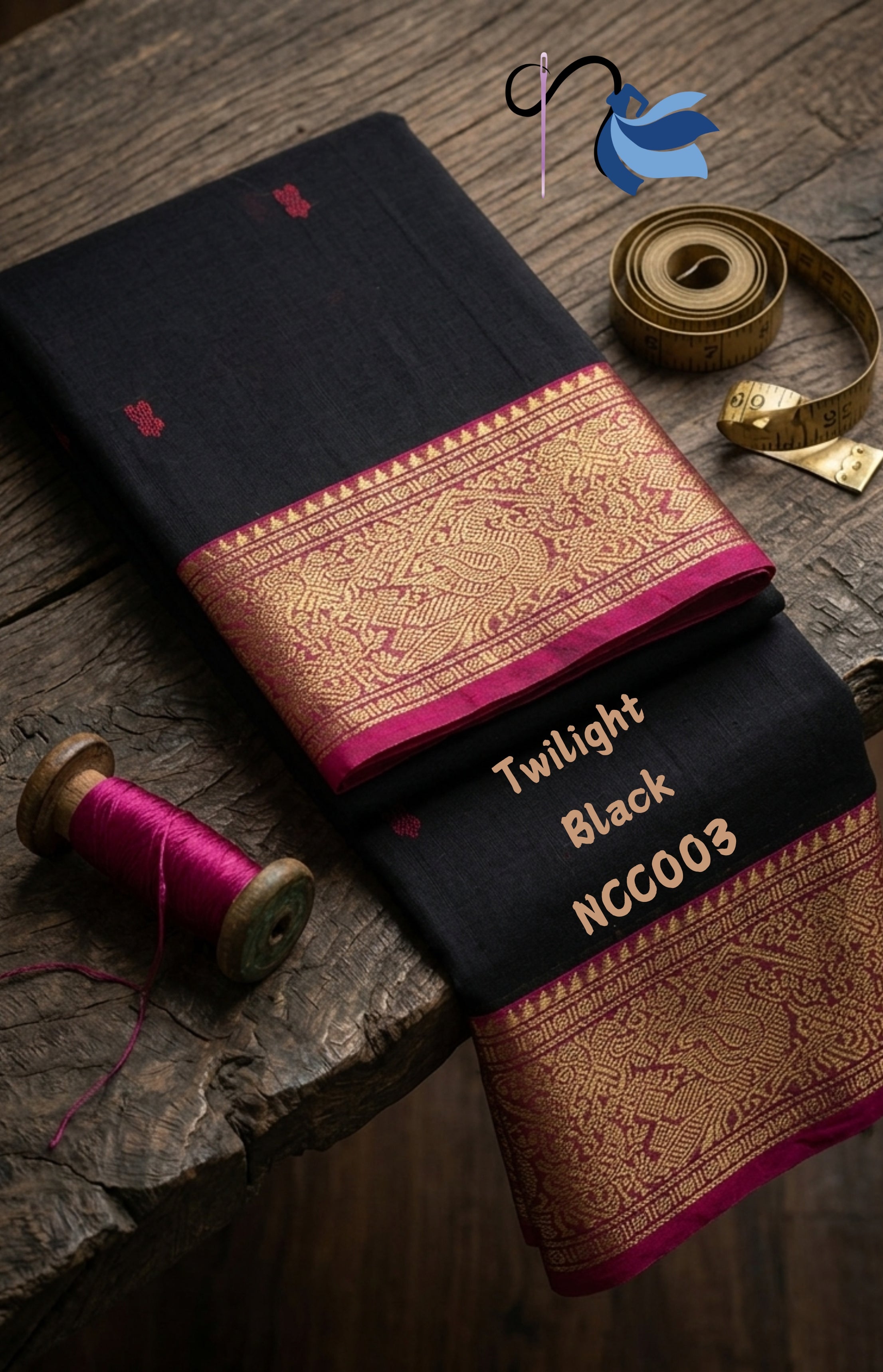 High-angle flat lay of Twilight Black Chettinad Cotton Saree by NAANAL. The black saree is folded on a rustic wooden table, showcasing a vibrant magenta and gold zari border. A wooden bobbin with magenta thread and a brass measuring tape are placed as props, emphasizing the hand-crafted quality. Product code NCC003 is visible on the fabric.